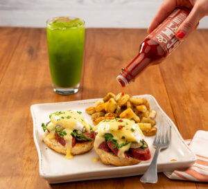 photo of person pouring Yi-Yi hot sauce on an eggs benedict with home style potatoes and a green juice in the background on a warm wood table