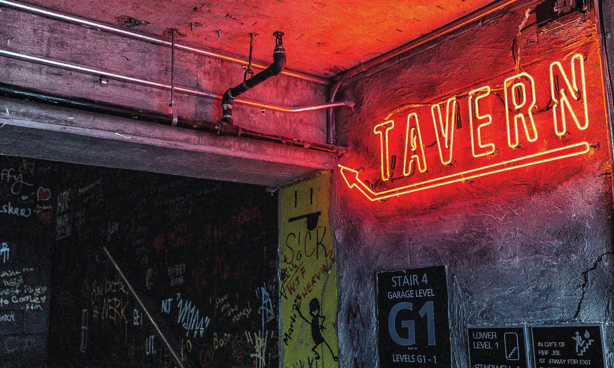 Red neon signage that reads TAVERN with an arrow pointing upstairs in a concrete garage