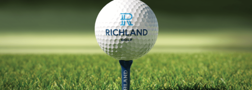 Image shows a golf ball on on a tee with blurred grassy background, on the golf ball is the Richland Country Club main logo with the lettering mark on the tee