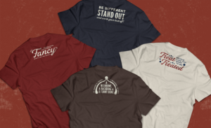 Reba's Place T-Shirt designs laid in flat lay. From left to right: Red shirt with "Here's your one chance Fancy don't let me down" , Blue shirt with "Be different, stand out, and work your but off!" , Brown shirt with "It takes 3 things to succeed in life: a wishbone, a backbone & a funny bone" , White shirt with "Treat people like you want to be treated"