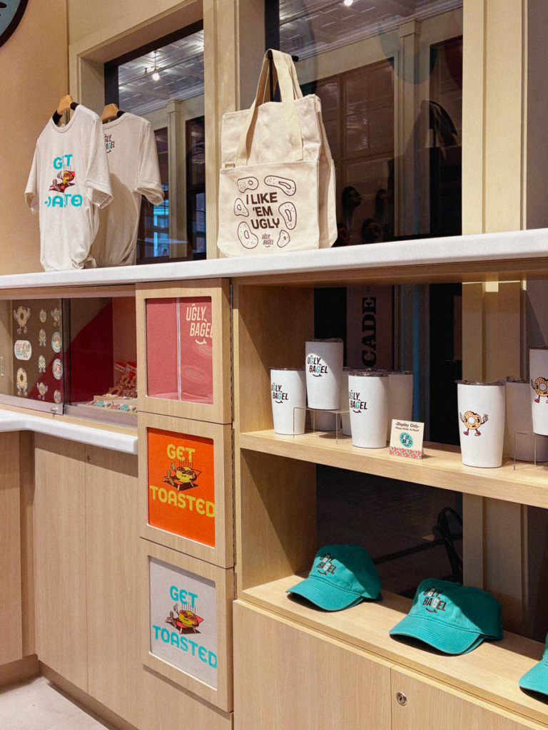 View of Ugly Bagel merch corner shelves with hats, shirts, tumblers, and tote bags